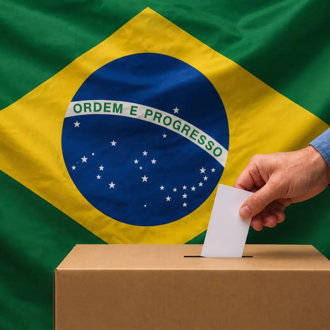 Mão depositando um voto em uma urna, com a bandeira do Brasil ao fundo.